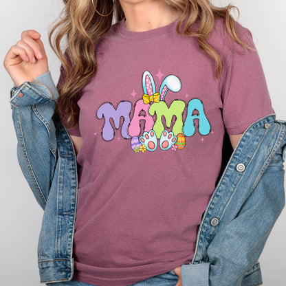 May include: A mauve-colored t-shirt with the word "MAMA" in colorful, bubble letters. The design includes bunny ears, Easter eggs, and bunny feet, creating a festive Easter theme. The shirt is a short-sleeved crew neck.
