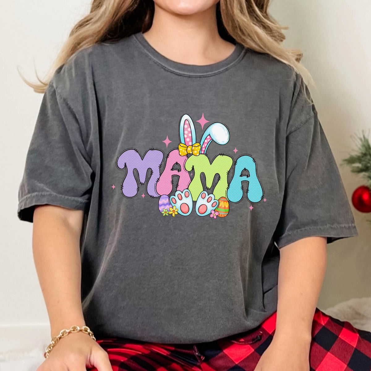 May include: Dark gray t-shirt with a colorful Easter-themed graphic. The word "MAMA" is displayed in bubble letters, with bunny ears and feet, and Easter eggs. The shirt is a relaxed fit.