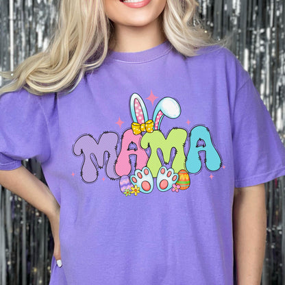 May include: A purple t-shirt with the word "MAMA" in a colorful, bubble-style font. The design includes bunny ears, Easter eggs, and paw prints, creating a festive Easter theme.
