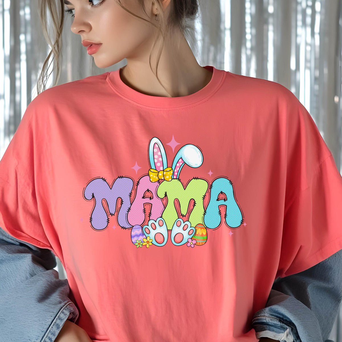 May include: Coral-colored t-shirt with the word "MAMA" in a colorful, cartoonish font. The design includes bunny ears, a bow, Easter eggs, and bunny feet, perfect for Easter or Mother's Day.