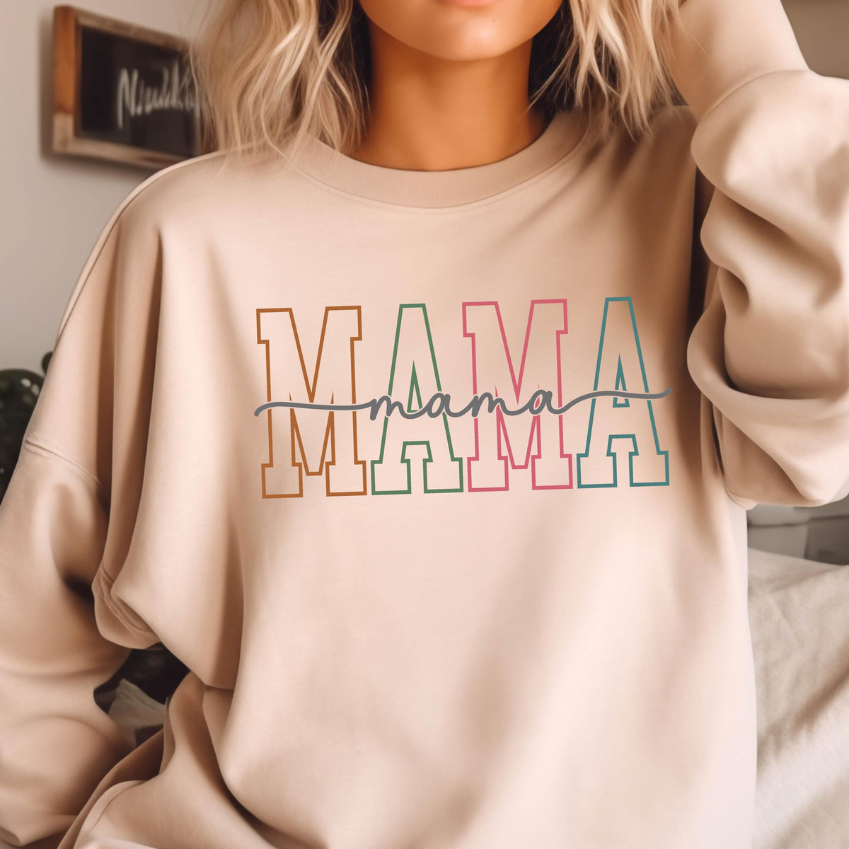 May include: A beige sweatshirt with the word "MAMA" printed in colorful block letters with a handwritten "mama" underneath.