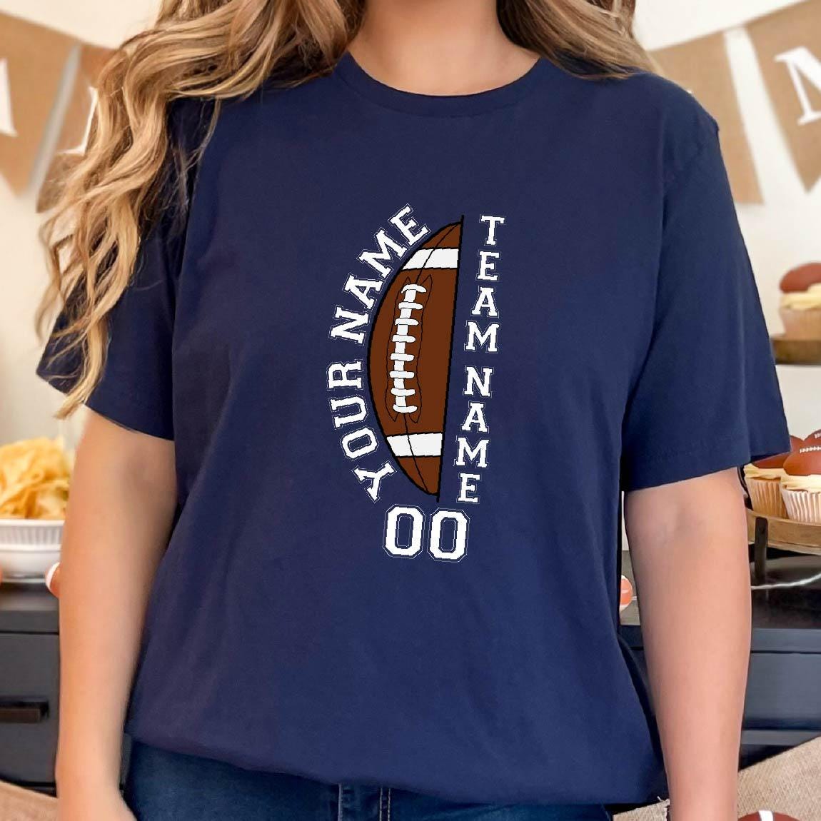 May include: A navy blue short-sleeve t-shirt featuring a graphic of a brown football. The text "YOUR NAME" is printed vertically on the left, "TEAM NAME" on the right, and the number "00" is centered below the football in white lettering.