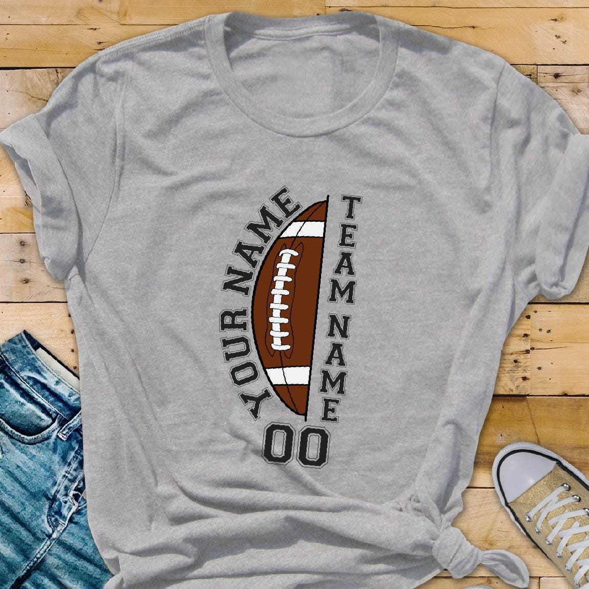 May include: A light gray short-sleeved t-shirt featuring a graphic of a brown football with white laces. The text "YOUR NAME" and "TEAM NAME" is printed vertically along the sides of the football, with the number "00" displayed below it.