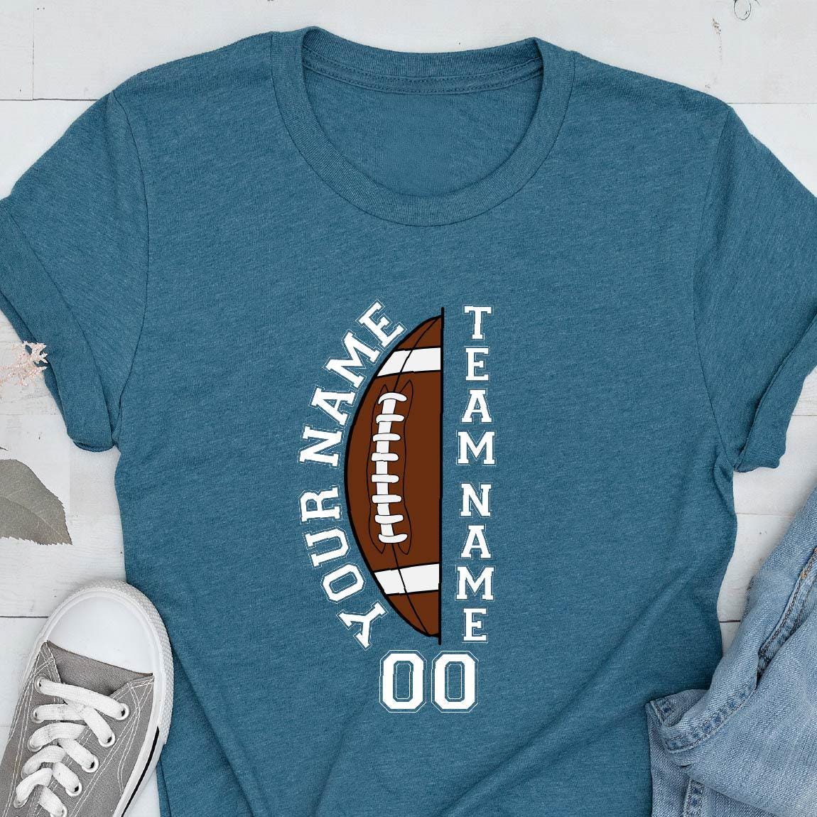 May include: A heathered teal crew neck t-shirt featuring a graphic of a brown football split in half. The text "YOUR NAME" is printed vertically on the left, "TEAM NAME" on the right, and the number "00" is centered below the football graphic.