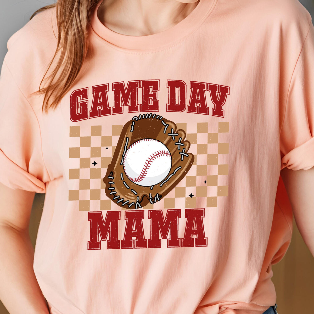May include: A peach-colored t-shirt featuring a graphic print with the text GAME DAY MAMA in red block letters. The design includes a brown baseball glove holding a white baseball with red stitching, set against a tan and brown checkered background.