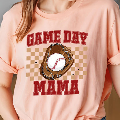 May include: A peach-colored t-shirt featuring a graphic print with the text GAME DAY MAMA in red block letters. The design includes a brown baseball glove holding a white baseball with red stitching, set against a tan and brown checkered background.
