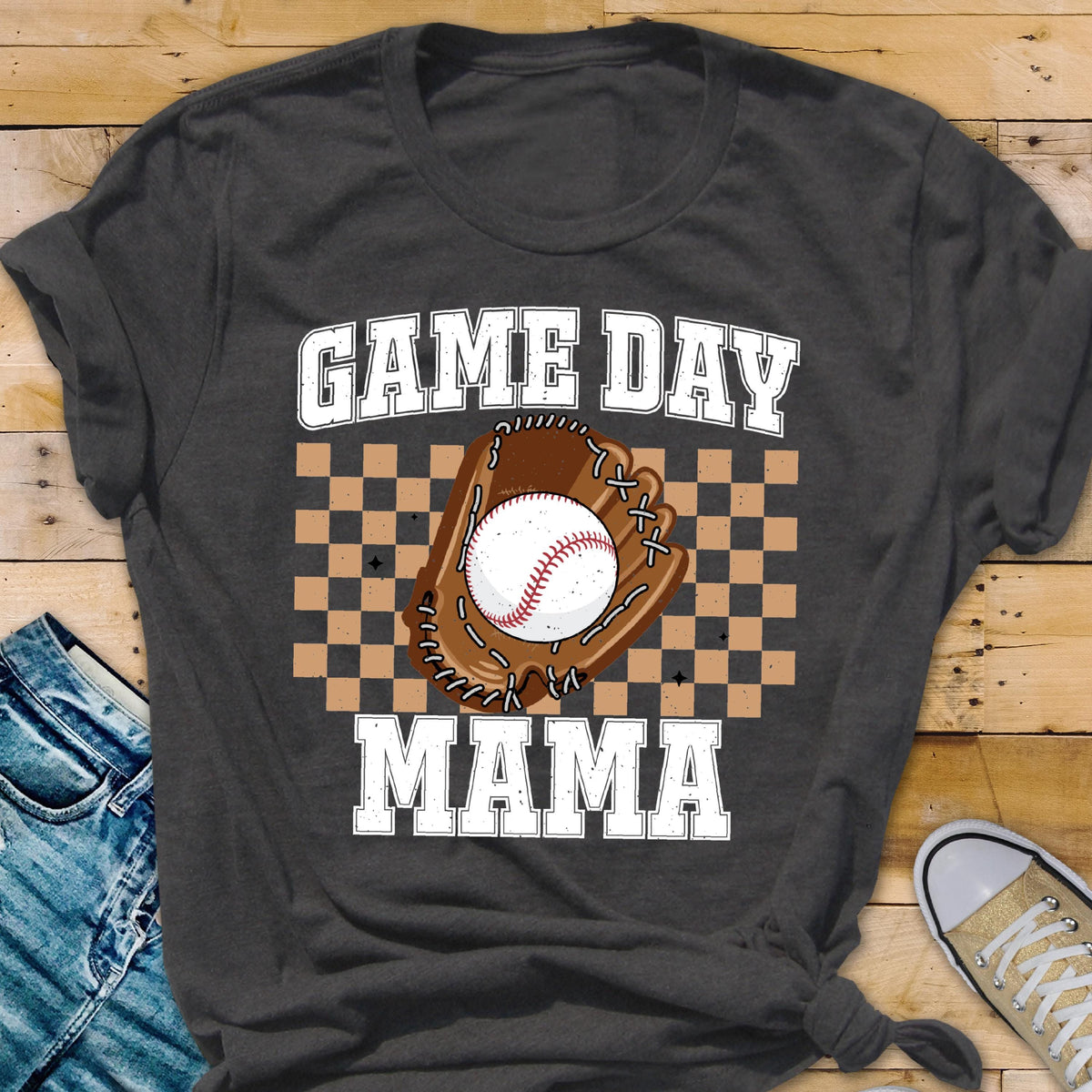 May include: A dark charcoal gray t-shirt featuring a graphic print with the text GAME DAY MAMA in white block letters. The design includes a brown baseball glove holding a white baseball, centered over a tan and dark brown checkered background.