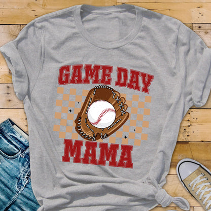 May include: A light gray short-sleeve t-shirt featuring a graphic print with the text GAME DAY MAMA in red block letters. The design includes a brown baseball glove holding a white baseball, centered over a tan checkered pattern background.