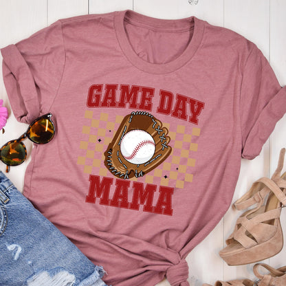 May include: A dusty rose short-sleeved t-shirt featuring a graphic print of a brown baseball glove holding a white baseball. The text "GAME DAY MAMA" is printed in red block letters above and below the glove, set against a tan checkered background.