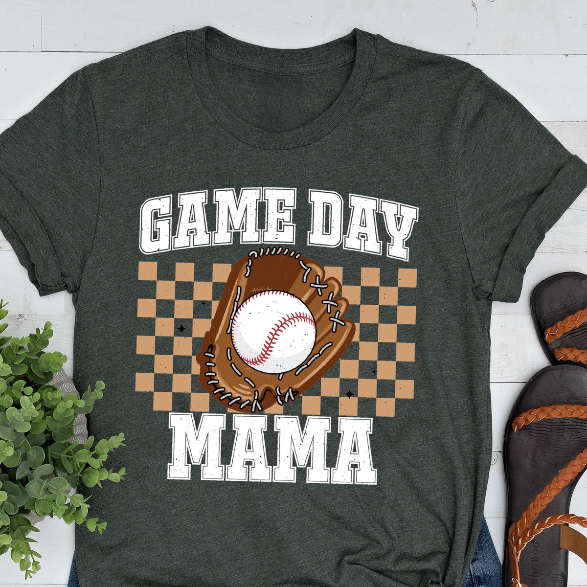 May include: A dark charcoal gray t-shirt featuring a graphic print with the text GAME DAY MAMA in white block letters. The center design includes a brown baseball glove holding a white baseball, set against a tan and brown checkered background.