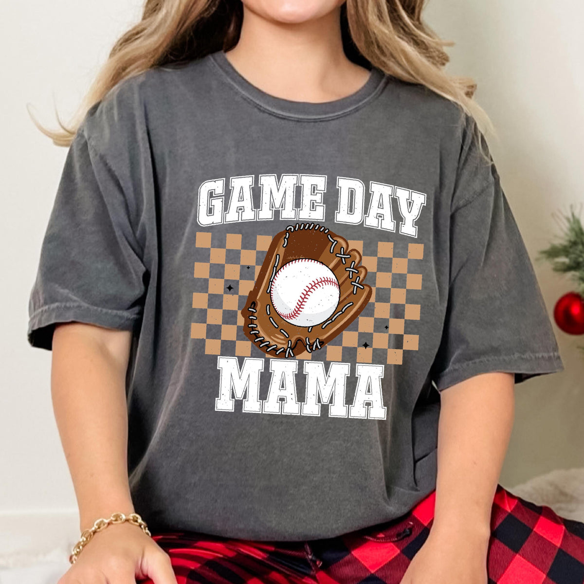 May include: A charcoal grey short-sleeve t-shirt featuring a graphic print with the text GAME DAY MAMA in white block letters. The design includes a brown baseball glove holding a white baseball, centered over a tan and brown checkered pattern.