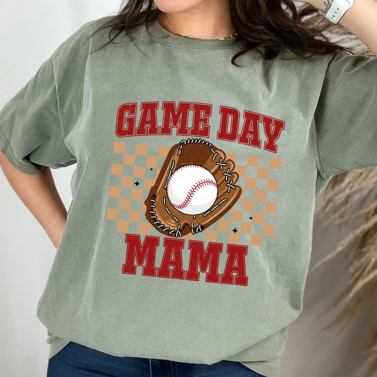 May include: A sage green short-sleeve t-shirt featuring a graphic print with the red text GAME DAY MAMA. The design includes a brown baseball glove holding a white baseball, centered over a tan checkered pattern with small black star accents.