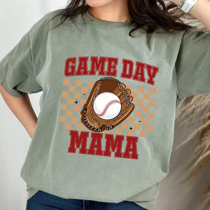 May include: A sage green short-sleeve t-shirt featuring a graphic print with the red text GAME DAY MAMA. The design includes a brown baseball glove holding a white baseball, centered over a tan checkered pattern with small black star accents.
