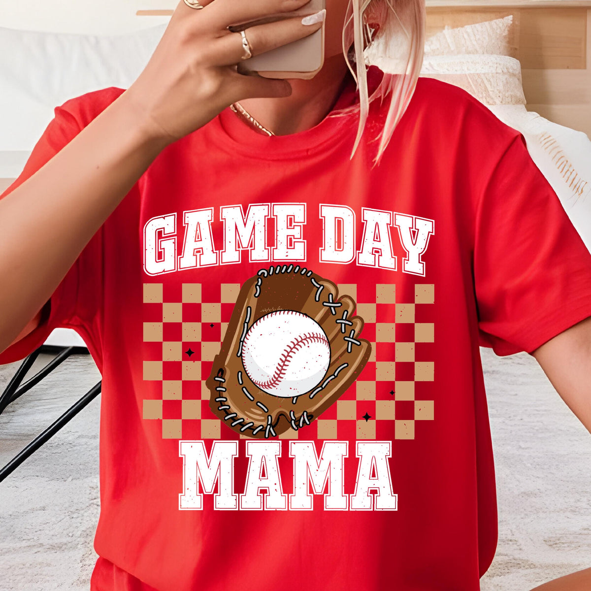 May include: A bright red short-sleeved t-shirt featuring a graphic print with the text GAME DAY MAMA in white block letters. Centered is a brown baseball glove holding a white baseball, set against a tan and brown checkered background pattern.