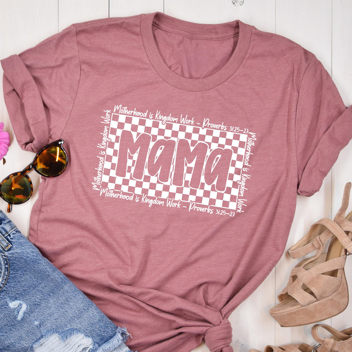 May include: A pink t-shirt with a white checkered background and the word "Mama" in white text. The text around the checkered background reads "Motherhood is Kingdom Work - Proverbs 31:25-27".
