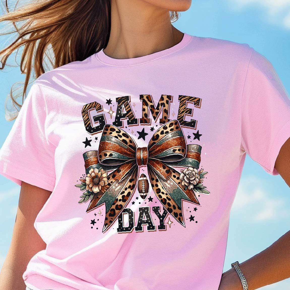 May include: Pink t-shirt with a graphic print of a leopard print bow with a football in the center. The text "GAME DAY" is printed above the bow.