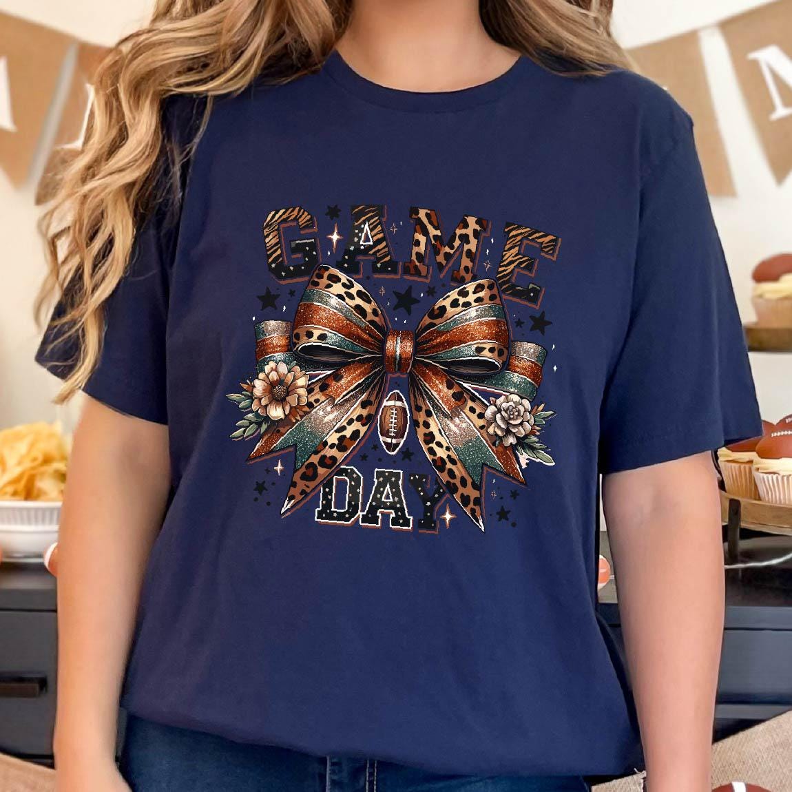 May include: A navy blue t-shirt with a graphic of a leopard print bow with a football in the center. The text "GAME DAY" is written in black letters above and below the bow.