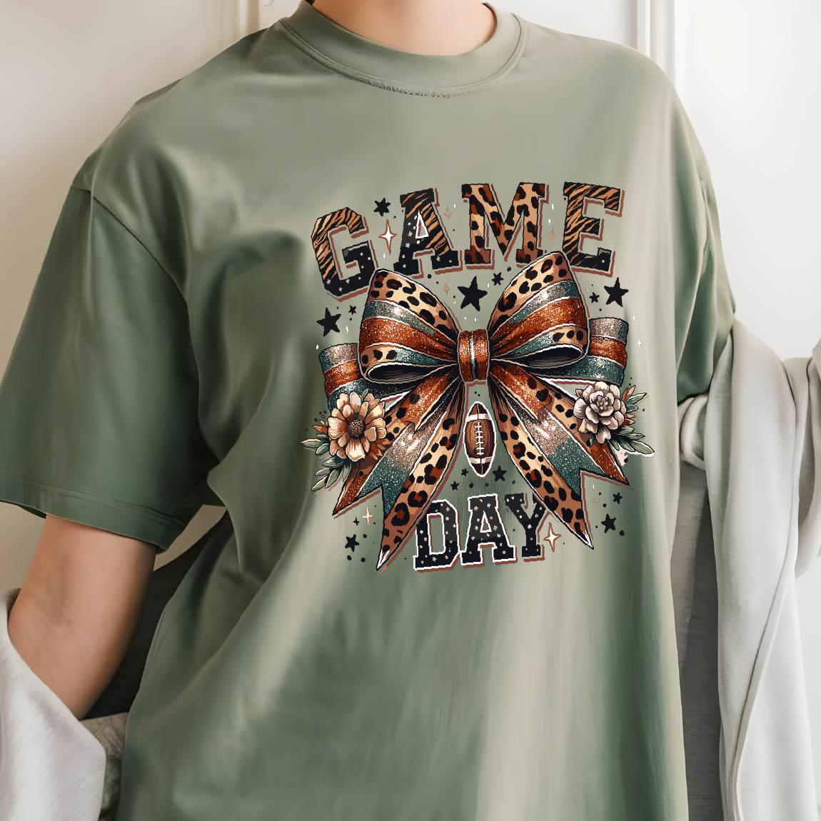 May include: A sage green t-shirt featuring a graphic print with the text "GAME DAY" in a leopard print pattern. The design includes a large central bow with leopard and glitter textures, small flowers, a football icon, and scattered stars.