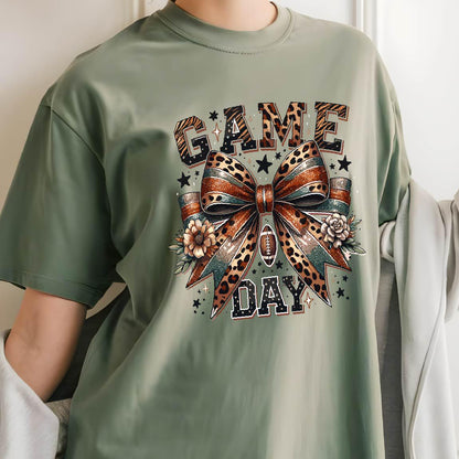 May include: A sage green t-shirt featuring a graphic print with the text "GAME DAY" in a leopard print pattern. The design includes a large central bow with leopard and glitter textures, small flowers, a football icon, and scattered stars.