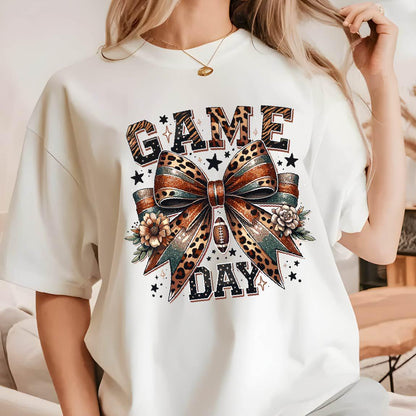 May include: A white short-sleeve t-shirt featuring a graphic print of a large bow with leopard and teal patterns, surrounded by flowers, stars, and a football. The text "GAME DAY" is printed in bold, dark letters above and below the bow design.