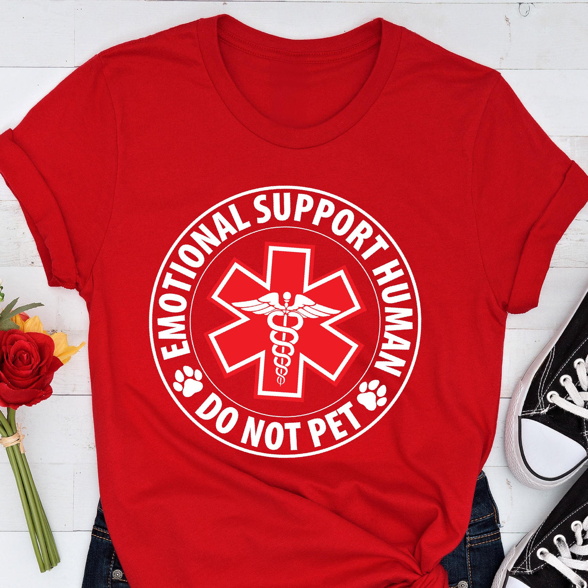May include: A red t-shirt featuring a white graphic with the text "EMOTIONAL SUPPORT HUMAN DO NOT PET". The graphic includes a medical symbol with a caduceus in the center and paw prints.