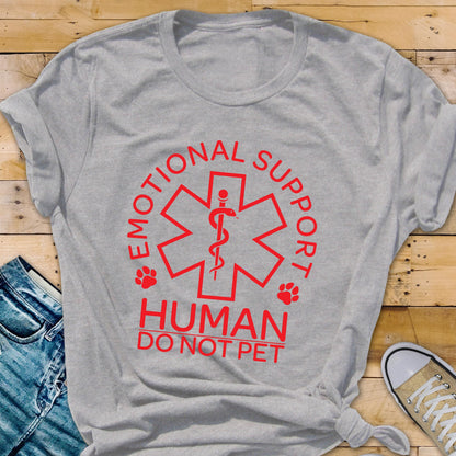 May include: A light gray short-sleeved t-shirt featuring a red graphic of a six-pointed star with a central staff and snake symbol. The text reads EMOTIONAL SUPPORT HUMAN DO NOT PET, accompanied by two small red paw print icons on either side.