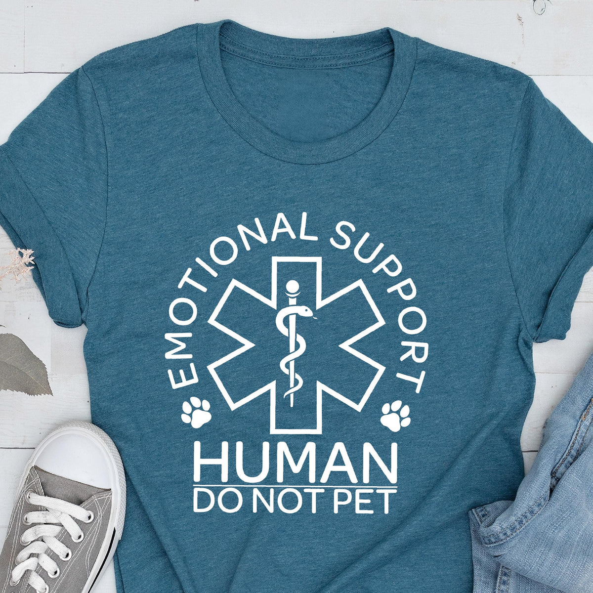 May include: A teal short-sleeved t-shirt featuring a white graphic of a six-pointed star of life symbol with a snake and staff. The text reads EMOTIONAL SUPPORT HUMAN DO NOT PET, accompanied by two small paw print icons on either side of the central emblem.