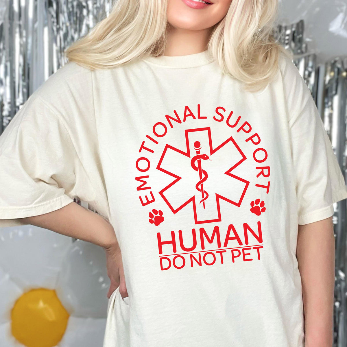 May include: An off-white short-sleeve t-shirt featuring a red graphic of a six-pointed star of life symbol with a snake and staff. The text reads EMOTIONAL SUPPORT HUMAN DO NOT PET, accompanied by two small red paw prints on either side of the star.