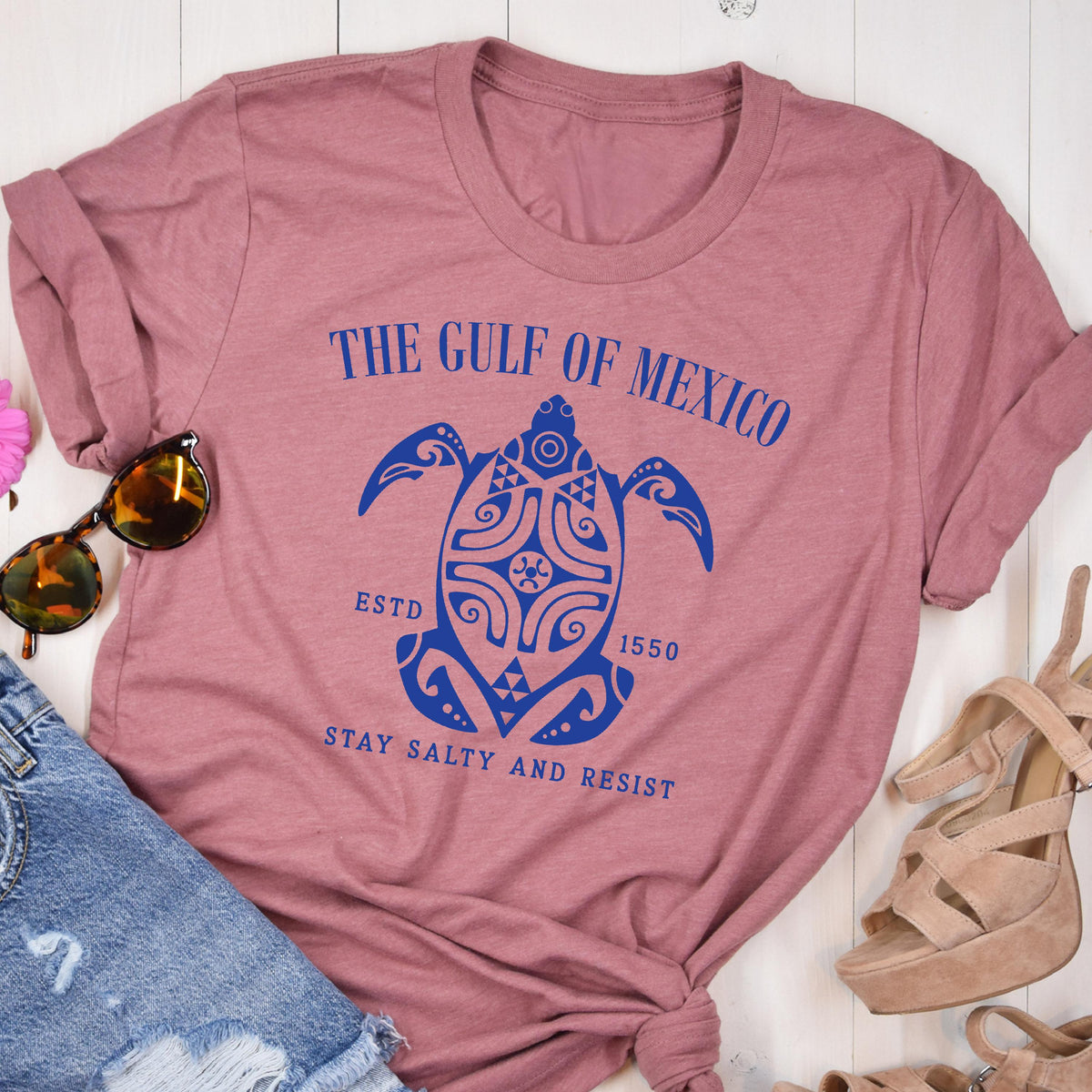 May include: A pink t-shirt with a blue graphic of a turtle with the text "The Gulf of Mexico" above it, "Est. 1550" below it, and "Stay Salty and Resist" below the turtle.