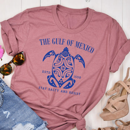 May include: A pink t-shirt with a blue graphic of a turtle with the text "The Gulf of Mexico" above it, "Est. 1550" below it, and "Stay Salty and Resist" below the turtle.