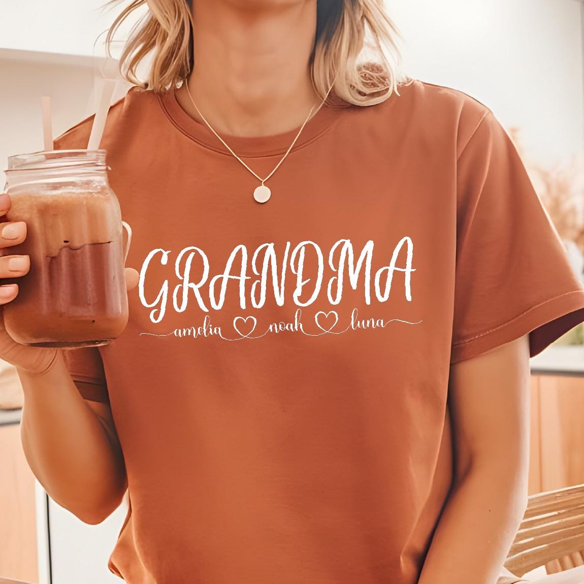 May include: A brown t-shirt with the text "Grandma" in white cursive lettering. Below the text are the names "Amelia", "Noah", and "Luna" with a heart symbol between each name.