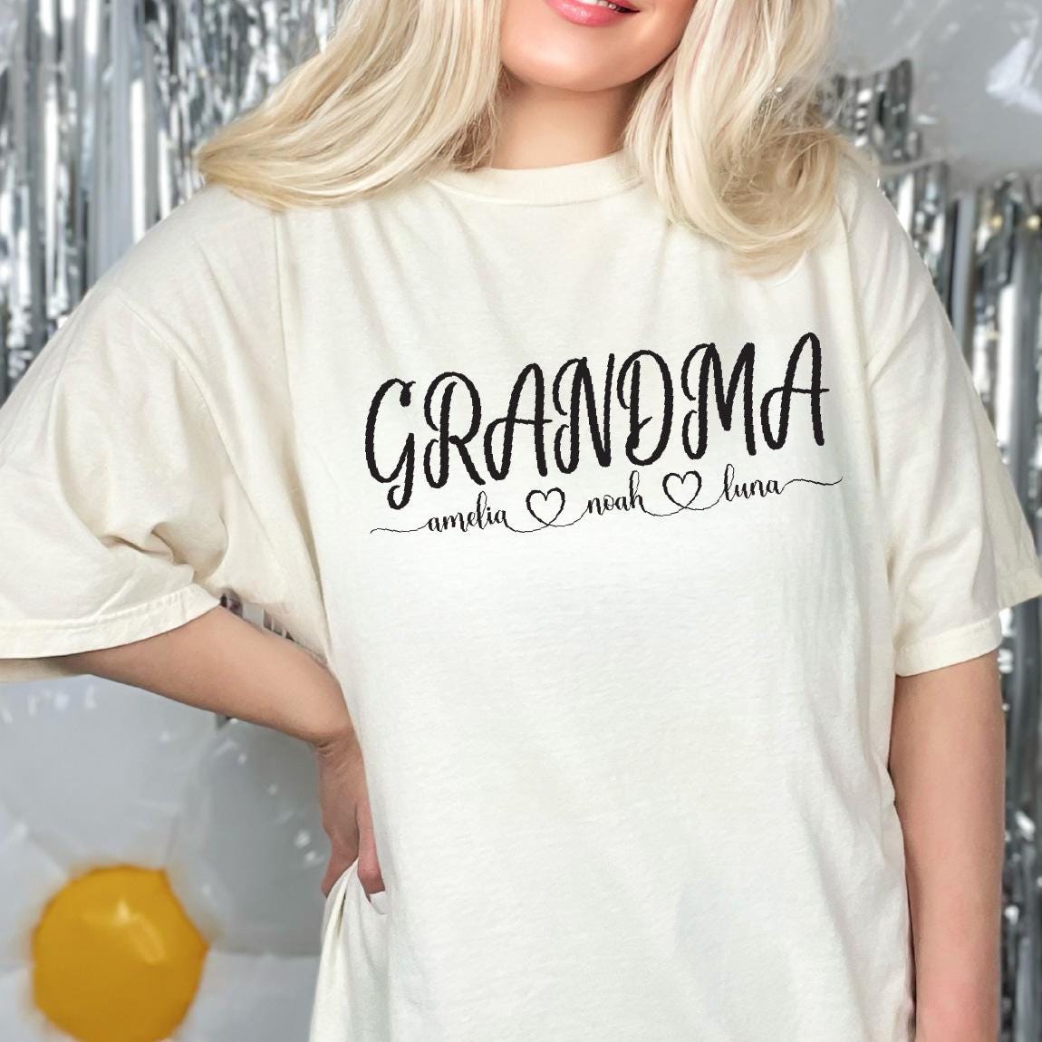 May include: White t-shirt with the text "Grandma" in black cursive lettering. Below the text are three names, "Amelia", "Noah", and "Luna", each followed by a small heart.