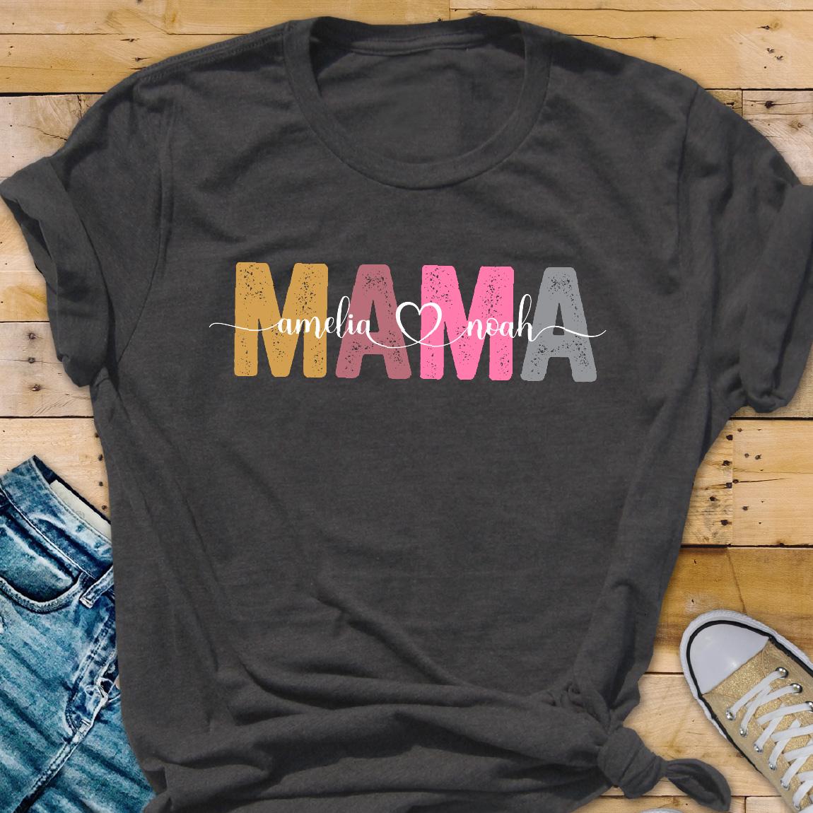 May include: A dark grey t-shirt with the word "MAMA" in colorful letters. The letters are in a distressed font and have a heart between the "M" and "A". The text "amelia" and "noah" are written in a smaller font below the word "MAMA".
