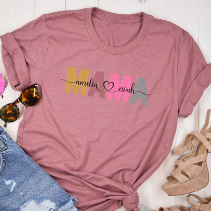 May include: A pink t-shirt with the word "MAMA" in colorful letters. The names "amelia" and "noah" are written in cursive script on either side of the word "MAMA" with a heart symbol between the names.