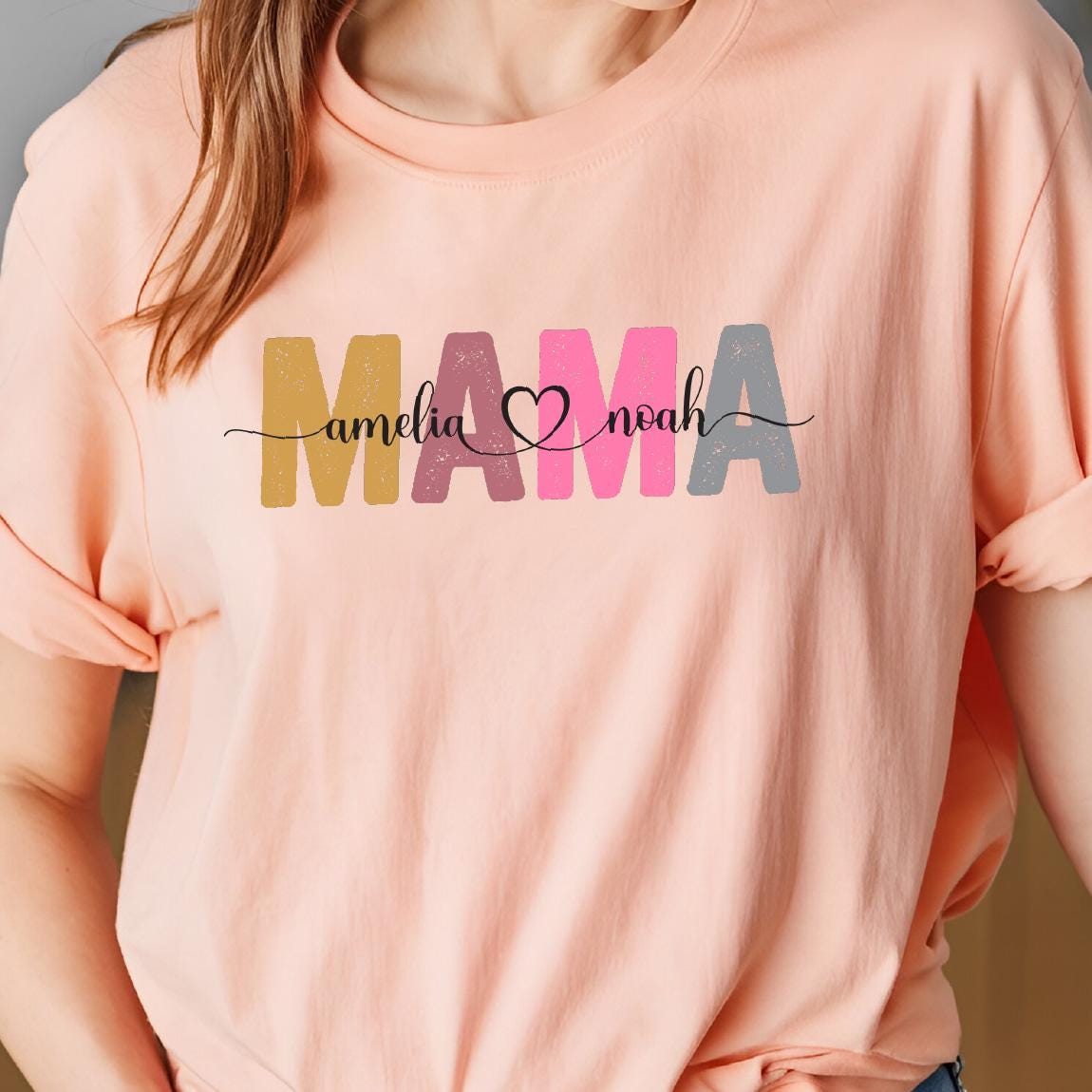 May include: A pink t-shirt with the word "MAMA" in colorful letters. The letters are decorated with a distressed, vintage look. The names "amelia" and "noah" are written in cursive on either side of the word "MAMA" with a heart symbol between the names.