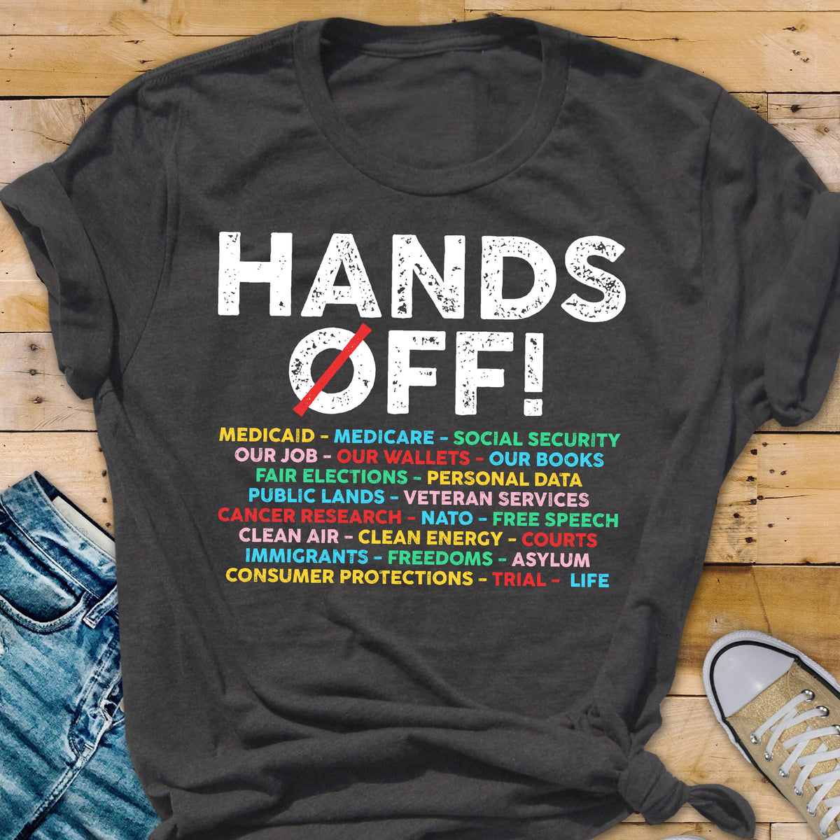 May include: A dark gray t-shirt with the text "HANDS OFF!" in white with a red slash through a circle. Below the text is a list of items in various colors, including "Medicaid", "Medicare", "Social Security", "Our Job", "Our Wallets", "Our Books", "Fair Elections", "Personal Data", "Public Lands", "Veteran Services", "Cancer Research", "NATO", "Free Speech", "Clean Air", "Clean Energy", "Courts", "Immigrants", "Freedoms", "Asylum", "Consumer Protections", "Trial", and "Life".