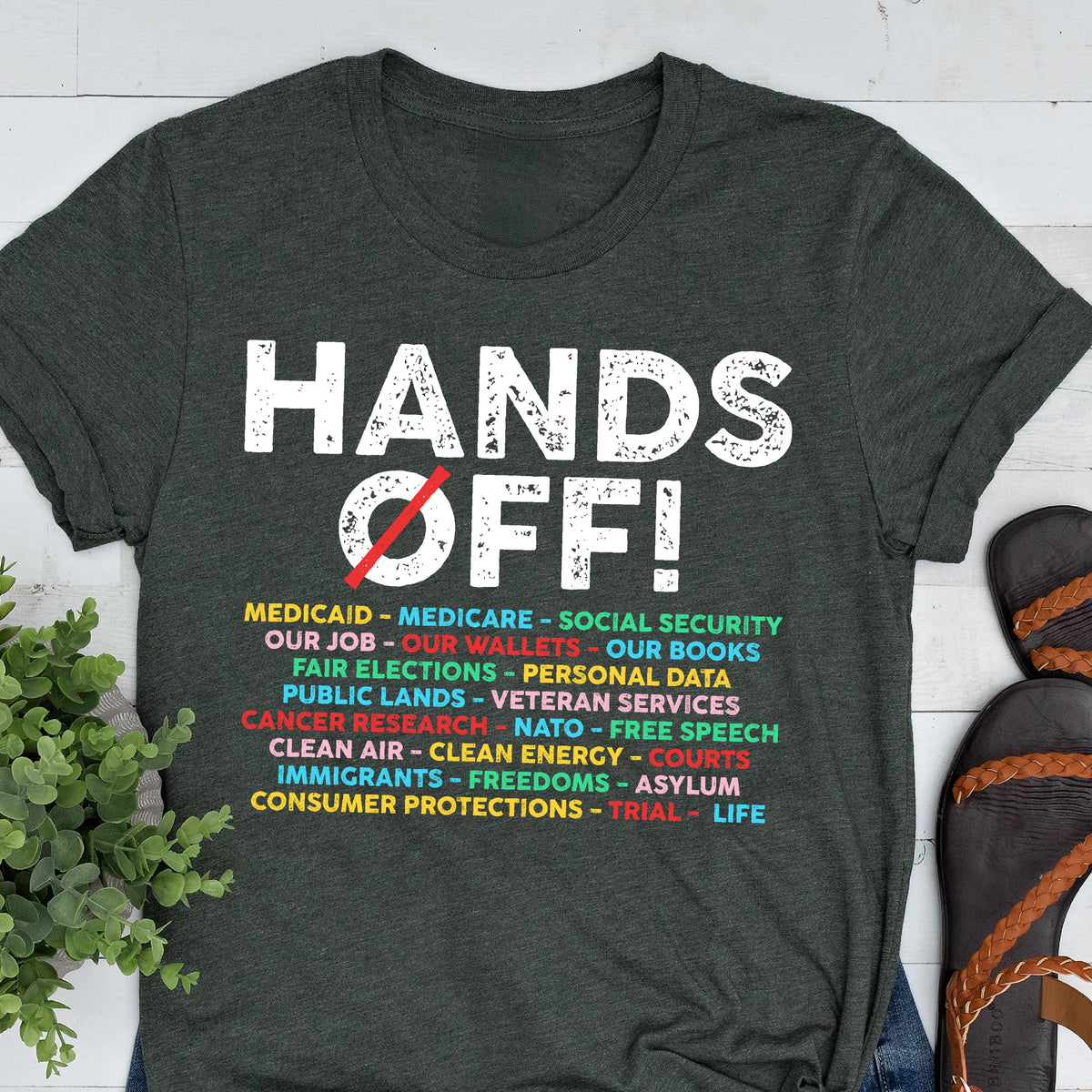 May include: A dark green t-shirt with the text "HANDS OFF!" in white. Below the text is a list of items in colorful text, including "Medicaid", "Medicare", "Social Security", "Our Job", "Our Wallets", "Our Books", "Fair Elections", "Personal Data", "Public Lands", "Veteran Services", "Cancer Research", "NATO", "Free Speech", "Clean Air", "Clean Energy", "Courts", "Immigrants", "Freedoms", "Asylum", "Consumer Protections", "Trial", and "Life".