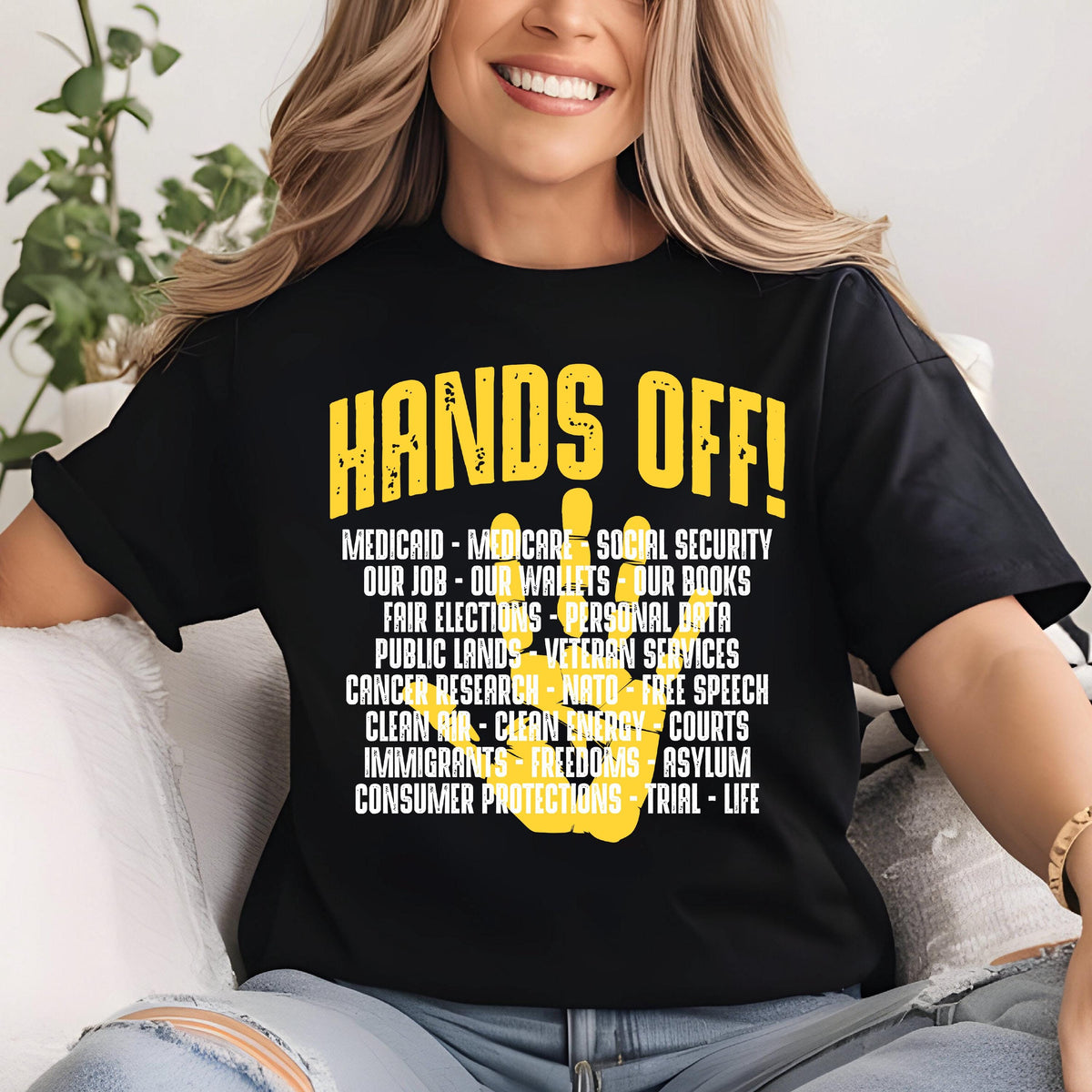 May include: Black t-shirt with a yellow handprint and the text "HANDS OFF!"  The text below the handprint lists various social and political issues, including Medicaid, Medicare, Social Security, our jobs, our wallets, our books, fair elections, personal data, public lands, veteran services, cancer research, NATO, free speech, clean air, clean energy, courts, immigrants, freedoms, asylum, consumer protections, trial, and life.