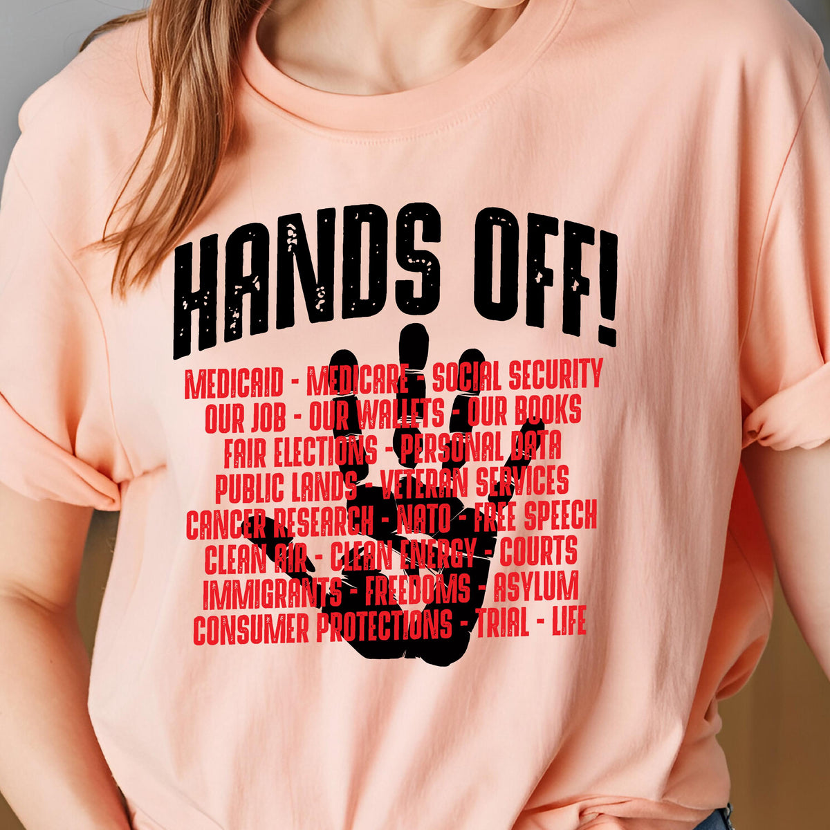 May include: A peach-colored t-shirt with the text "HANDS OFF!" in black. The text is surrounded by a black handprint with the following text in red: "MEDICAID - MEDICARE - SOCIAL SECURITY - OUR JOB - OUR WALLETS - OUR BOOKS - FAIR ELECTIONS - PERSONAL DATA - PUBLIC LANDS - VETERAN SERVICES - CANCER RESEARCH - NATO - FREE SPEECH - CLEAN AIR - CLEAN ENERGY - COURTS - IMMIGRANTS - FREEDOMS - ASYLUM - CONSUMER PROTECTIONS - TRIAL - LIFE"
