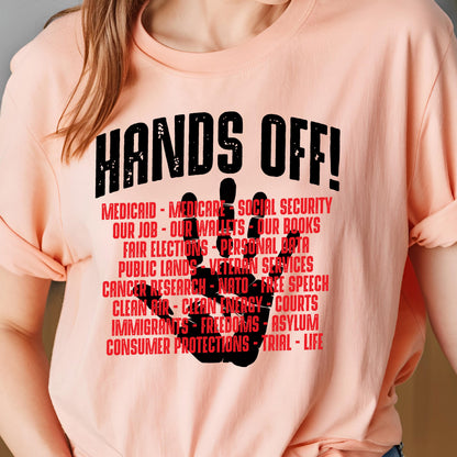 May include: A peach-colored t-shirt with the text "HANDS OFF!" in black. The text is surrounded by a black handprint with the following text in red: "MEDICAID - MEDICARE - SOCIAL SECURITY - OUR JOB - OUR WALLETS - OUR BOOKS - FAIR ELECTIONS - PERSONAL DATA - PUBLIC LANDS - VETERAN SERVICES - CANCER RESEARCH - NATO - FREE SPEECH - CLEAN AIR - CLEAN ENERGY - COURTS - IMMIGRANTS - FREEDOMS - ASYLUM - CONSUMER PROTECTIONS - TRIAL - LIFE"