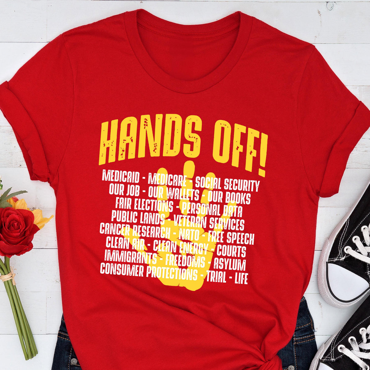 May include: Red t-shirt with a yellow handprint and the text "HANDS OFF!" with a list of items including "Medicaid", "Medicare", "Social Security", "Our Job", "Our Wallets", "Our Books", "Fair Elections", "Personal Data", "Public Lands", "Veteran Services", "Cancer Research", "NATO", "Free Speech", "Clean Air", "Clean Energy", "Courts", "Immigrants", "Freedoms", "Asylum", "Consumer Protections", "Trial", and "Life".