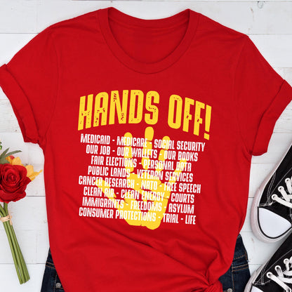 May include: Red t-shirt with a yellow handprint and the text "HANDS OFF!" with a list of items including "Medicaid", "Medicare", "Social Security", "Our Job", "Our Wallets", "Our Books", "Fair Elections", "Personal Data", "Public Lands", "Veteran Services", "Cancer Research", "NATO", "Free Speech", "Clean Air", "Clean Energy", "Courts", "Immigrants", "Freedoms", "Asylum", "Consumer Protections", "Trial", and "Life".