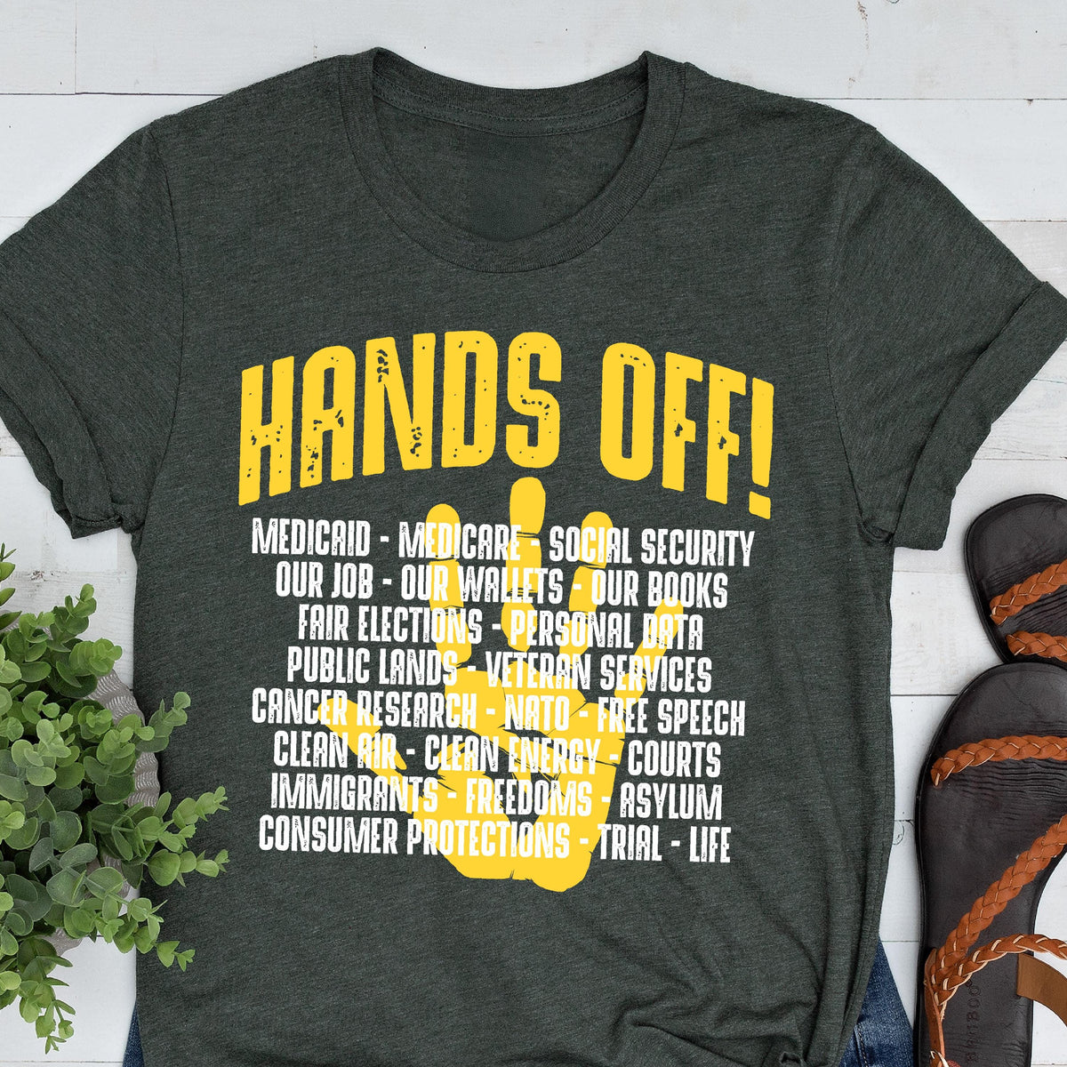 May include: A dark green t-shirt with a yellow peace sign and the text "HANDS OFF!"  The text below the peace sign lists various social and political issues, including "Medicaid", "Medicare", "Social Security", "Our Job", "Our Wallets", "Our Books", "Fair Elections", "Personal Data", "Public Lands", "Veteran Services", "Cancer Research", "NATO", "Free Speech", "Clean Air", "Clean Energy", "Courts", "Immigrants", "Freedoms", "Asylum", "Consumer Protections", "Trial", and "Life".