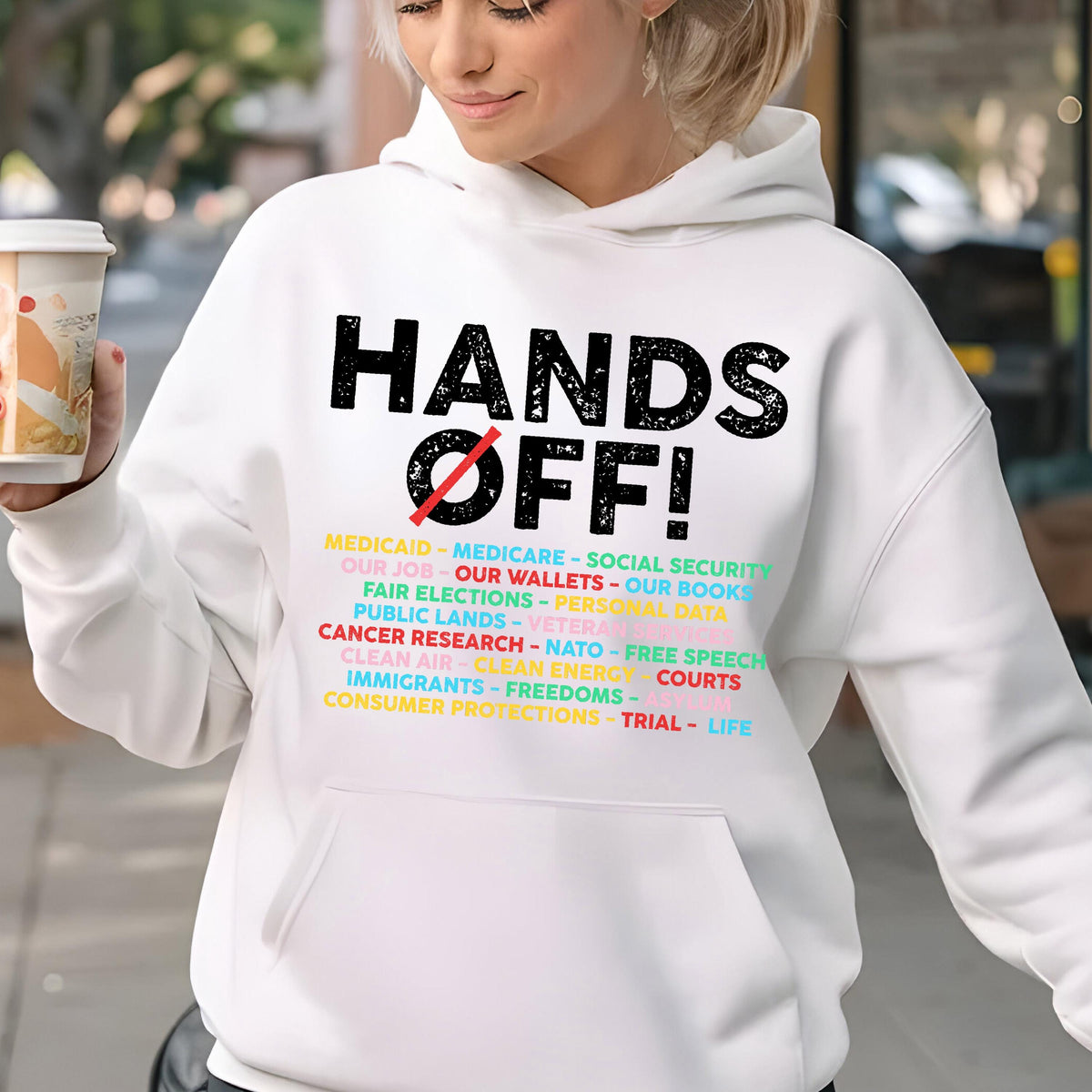 May include: White hoodie with a black graphic that says "Hands Off!" and a list of issues including "Medicaid", "Medicare", "Social Security", "Our Job", "Our Wallets", "Our Books", "Fair Elections", "Public Lands", "Veterans", "Cancer Research", "NATO", "Free Speech", "Clean Air", "Clean Energy", "Courts", "Immigrants", "Freedoms", "Trial", and "Life".