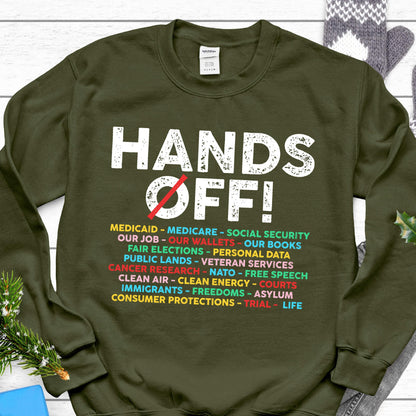 May include: Green sweatshirt with the text "HANDS OFF!" in white with a red slash through the O in OFF. Below the text is a list of items in various colors including: Medicaid, Medicare, Social Security, Our Job, Our Wallets, Our Books, Fair Elections, Personal Data, Public Lands, Veteran Services, Cancer Research, NATO, Free Speech, Clean Air, Clean Energy, Courts, Immigrants, Freedoms, Asylum, Consumer Protections, Trial, Life.