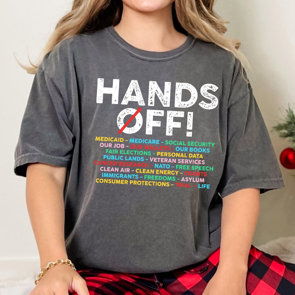 May include: A charcoal gray short-sleeve t-shirt featuring the white distressed text HANDS OFF! with a red diagonal line through the word OFF. Below, a list of topics in yellow, blue, and red text includes Medicaid, Medicare, and Social Security.