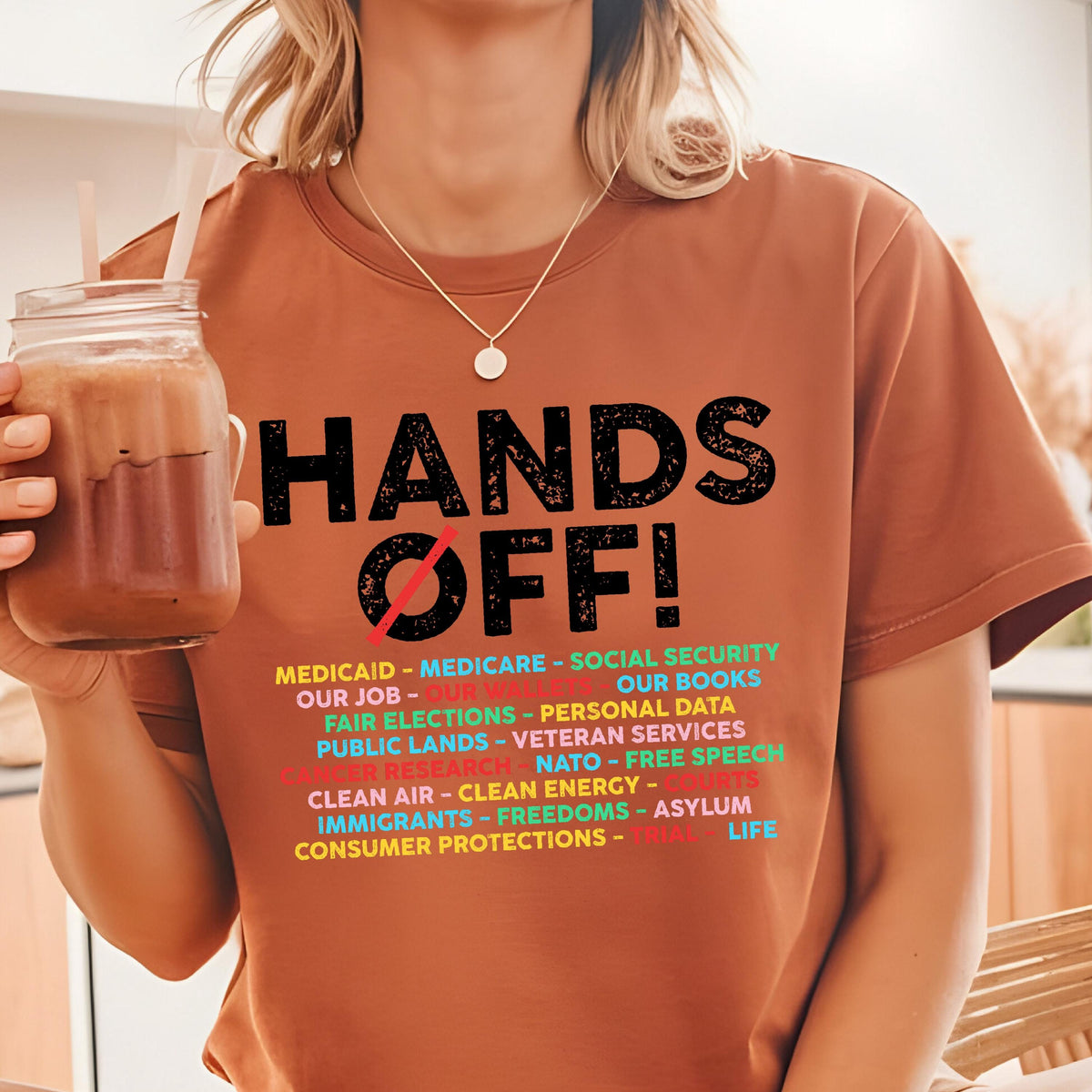 May include: A rust-colored t-shirt featuring the black text HANDS OFF! with a red slash through the O. Below, a list of multi-colored text includes Medicaid, Medicare, Social Security, Our Job, Our Wallet, Our Books, Fair Elections, and Personal Data.