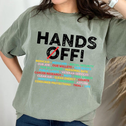May include: A sage green short-sleeve t-shirt featuring the black text HANDS OFF! with a red diagonal line through the O. Below, a list of multi-colored words includes Medicaid, Medicare, Social Security, Our Job, Our Wallets, Our Books, and more.
