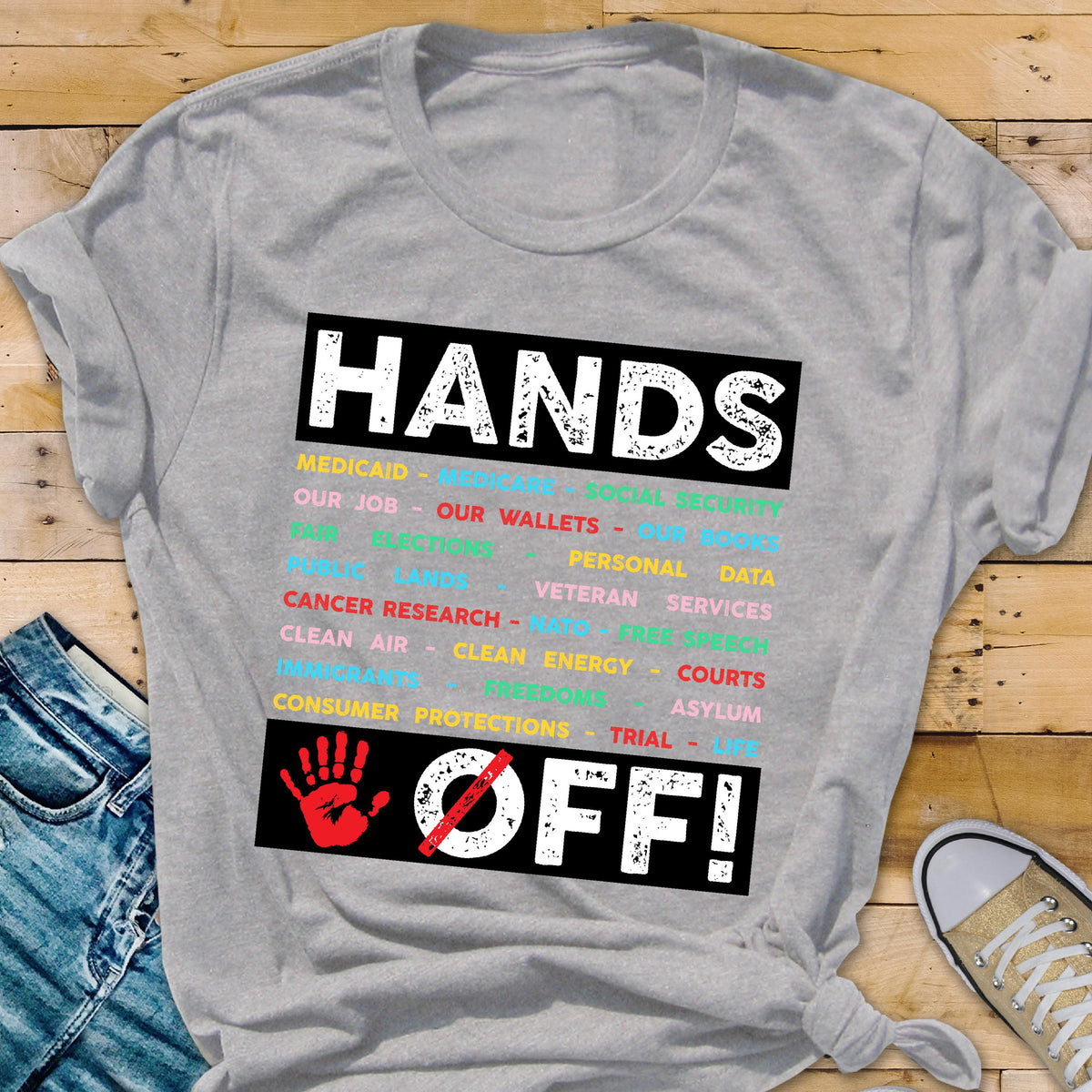 May include: Gray t-shirt with a black rectangle featuring the text "HANDS OFF!" in white with a red handprint and a diagonal red line through it. The rectangle is filled with a list of words in various colors, including "Medicaid", "Our Job", "Our Wallets", "Personal Data", "Veteran Services", "Cancer Research", "Clean Air", "Clean Energy", "Courts", "Asylum", "Trial", and "Consumer Protections".
