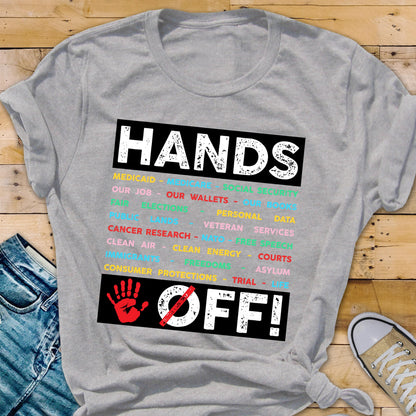 May include: Gray t-shirt with a black rectangle featuring the text "HANDS OFF!" in white with a red handprint and a diagonal red line through it. The rectangle is filled with a list of words in various colors, including "Medicaid", "Our Job", "Our Wallets", "Personal Data", "Veteran Services", "Cancer Research", "Clean Air", "Clean Energy", "Courts", "Asylum", "Trial", and "Consumer Protections".